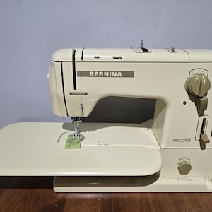 May include: Cream-colored Bernina Record sewing machine. The machine has a large, flat extension table and several dials and levers. The word "BERNINA" is printed on the front, and "record" is printed on the side. The machine is on a wooden surface.