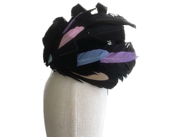 May include: A black feather fascinator with accents of pastel pink, blue, and lavender feathers. The headpiece is mounted on a mannequin head. The design includes the text "Anne New York DePasquale adhats".