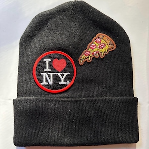 Peut inclure: Bonnet noir avec deux écussons brodés. L'un affiche "I NY" avec un cœur rouge, l'autre une part de pizza au pepperoni et au fromage. Un accessoire d'hiver classique.