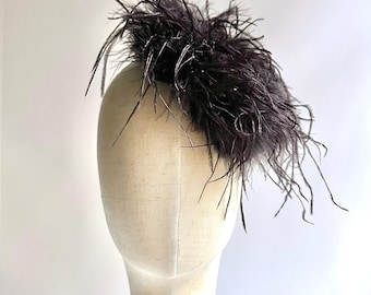 May include: A dark gray feather fascinator displayed on a mannequin head. The headpiece features a dense cluster of dark gray feathers, with some extending outwards. The mannequin is mounted on a gold-colored stand against a white background.