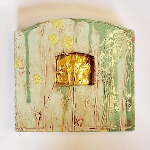 Op de afbeelding: Vierkante keramische wandkunst met een getextureerd oppervlak. Het kunstwerk heeft een goudkleurig metalen vierkant in het midden, omgeven door een lichtgroene en crèmekleurige achtergrond met bloemen en verticale lijnen. Het stuk heeft een afgeronde bovenrand.