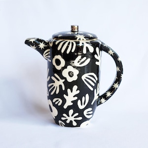 May include: Black and white ceramic teapot with a lid. The teapot is adorned with a repeating pattern of white floral and abstract shapes. The handle and spout are decorated with white star shapes. The lid has a silver-colored knob.