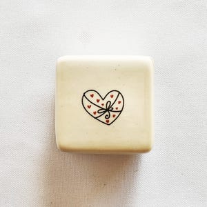 Peut inclure: Un petit plat carré en céramique blanc cassé. Un contour noir d'un coffret cadeau en forme de cœur est centré sur le plat, avec de petits cœurs rouges et un nœud. Le plat est un objet décoratif.