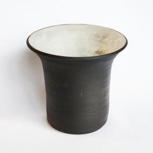 May include: A handcrafted, dark gray ceramic vase with a flared rim and a contrasting off-white interior. The vase has a cylindrical body and a smooth, matte finish. The design is simple and modern, suitable for displaying flowers or as a decorative piece.