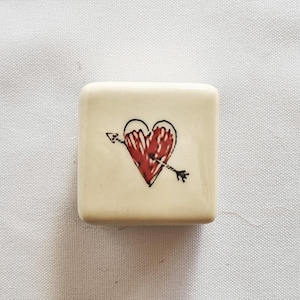 Puede incluir: Una baldosa cuadrada de cerámica blanquecina con un corazón rojo atravesado por una flecha negra. El corazón tiene una apariencia texturizada, y la flecha es negra con un extremo emplumado. La baldosa es probablemente un elemento decorativo.