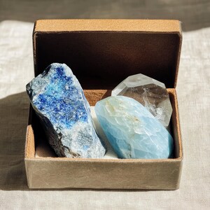 Puede incluir: Una caja marrón con la tapa abierta, revelando tres cristales. Los cristales son un lapislázuli azul, un cuarzo transparente y una aguamarina azul claro.