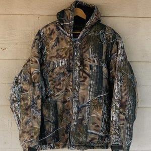 May include: A camouflage jacket with a hood, hanging against a light-colored wooden wall. The jacket features a full zipper closure and a drawstring at the bottom. The pattern includes shades of brown, green, and black, creating a woodland camouflage design.