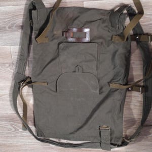 May include: Olive green canvas backpack with adjustable straps and multiple buckles. Features a front pocket with a flap and a leather patch. The bag is designed with a rectangular shape and is likely used for carrying items.