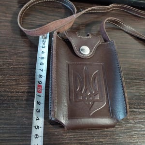 Może przedstawiać: Brązowa skórzana torba na ramię z paskiem, z ukraińskim emblematem trójzębu. Torba ma szerokość około 10 cm i posiada zapięcie na zatrzask. Torba leży na drewnianej powierzchni.