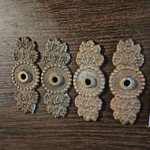May include: Four antique bronze drawer pulls with ornate floral and beaded designs. Each pull has a central hole and a decorative top and bottom. A ruler is next to the pulls, showing measurements in inches.