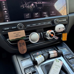 以下が含まれることがあります： 黒いダッシュボードと「設定」という文字が表示された画面のある車のインテリア。ダッシュボードには、「落ち着いて続けよう」という文字のノブや、「24」という数字と「コービー・ブライアント」という文字の書かれた木製のプレートなど、いくつかのボタンとノブがあります。また、ダッシュボードには、小さなエッセンシャルオイルのボトルがいくつかあります。