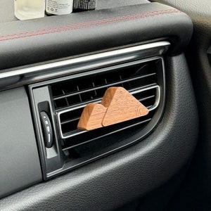 Peut inclure: Un diffuseur d'air de voiture en bois en forme de montagnes, avec une finition en bois brun et naturel. Le diffuseur est placé dans l'aérateur du tableau de bord d'une voiture. Trois petits flacons d'huiles essentielles sont posés sur le tableau de bord.
