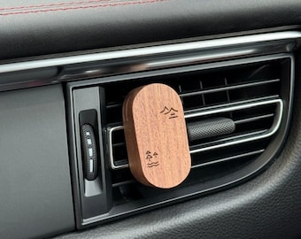 May include: A wooden car air freshener with a tree and water design. The air freshener is attached to a car's air vent. Two small bottles of essential oil are on the dashboard.