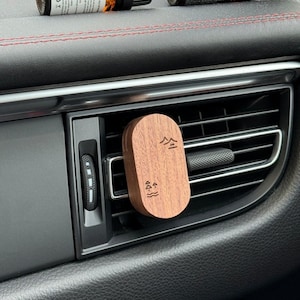 Puede incluir: Un ambientador de coche de madera con un diseño de árbol y agua. El ambientador está sujeto a una rejilla de ventilación del coche. Dos pequeñas botellas de aceite esencial están en el salpicadero.