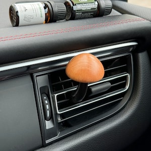 Könnte beinhalten: Ein Holz-Auto-Lüftungsclip mit einem braunen Holz-Knopf. Zwei kleine Glasflaschen mit ätherischem Öl befinden sich auf dem Armaturenbrett. Die Flaschen sind mit "Sandalwood" und "Cedar" beschriftet. Das Etikett der Zedernflasche lautet "Organic Plant & Natural 100% Pure essential oil 10ml (33)"