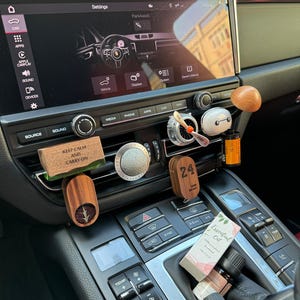 May include: A car's interior with a black dashboard and a touchscreen display. The display shows the car's settings menu. There are several buttons and knobs on the dashboard, including a temperature control panel. There are also several decorative items on the dashboard, including a wooden air freshener with the text "Keep Calm and Carry On", a wooden air freshener with the number "24", and a small bottle of essential oil.