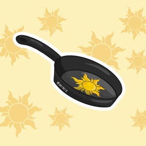 May include: A black frying pan with a white outline, featuring a yellow sun with rays in the center. The pan is surrounded by yellow suns with rays on a light yellow background.