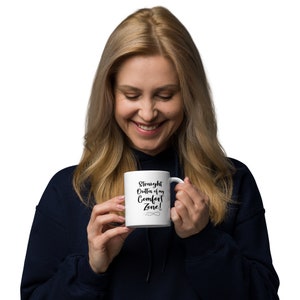 May include: A white ceramic mug with the text "Straight Outta of my Comfort Zone!" in black script. The mug has a curved handle and is held by a person wearing a navy blue hoodie.