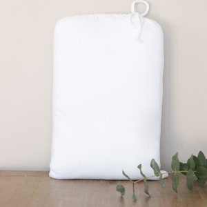 Peut inclure: Un oreiller blanc rectangulaire avec un dessus arrondi et une fermeture à cordon. L'oreiller est fait d'un tissu doux et lisse. Une branche de feuilles vertes se trouve dans le coin inférieur droit. L'oreiller est sur une surface en bois.