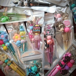 May include: Assortment of colorful beaded keychains with attached pens. The keychains feature various charms, including paw prints, hearts, and cartoon animals. The pens are decorated with phrases like "Live Love Meow" and "Kinda Sweet Kinda Savage".