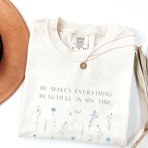 May include: Cream-colored t-shirt with the text "HE MAKES EVERYTHING BEAUTIFUL IN HIS TIME" and blue floral accents. A gold necklace with a pendant and a brown hat are also visible.