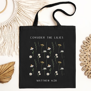 May include: Black tote bag with white text that reads "Consider the Lilies" and "Matthew 6:28". The bag features a pattern of white daisies and yellow flower centers on a black background.