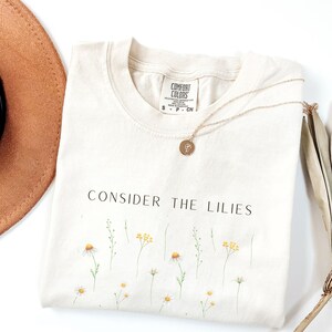 Puede incluir: Una camiseta de color crema con el texto "CONSIDER THE LILIES" impreso en negro. La camiseta presenta un pequeño patrón floral de margaritas blancas y centros amarillos.