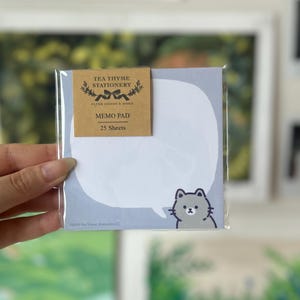 May include: A memo pad with a light blue background and a white speech bubble design. A gray cat illustration is on the right side. The packaging includes a brown label with the text "TEA THYME STATIONERY" and "MEMO PAD 25 Sheets."