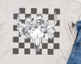 Peut inclure: Un t-shirt beige avec un motif à carreaux noir et blanc et un motif de crâne floral. Le crâne est entouré de fleurs et a un motif floral sur les cornes. Le texte "Wild Moon Designs" est visible sur le t-shirt.