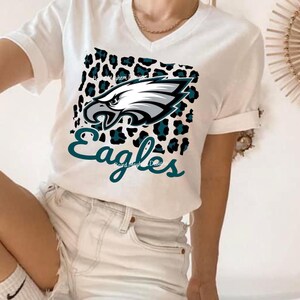 May include: White V-neck t-shirt with the Philadelphia Eagles logo. The logo features a silver and teal eagle head on a teal and black leopard print. The word "Eagles" is written in teal script below the logo. A casual, everyday garment.