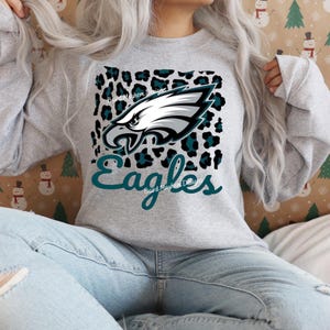 May include: A grey sweatshirt with the Philadelphia Eagles logo. The design features a white and teal eagle head, the word "Eagles" in teal script, and a leopard print background. The text "Baird Mayhem Designs" is also present.