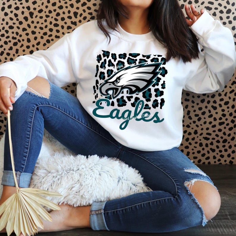 May include: White crew neck sweatshirt with the Philadelphia Eagles logo and a teal and black leopard print design. The word "Eagles" is written in teal script below the logo. The sweatshirt is a casual style.