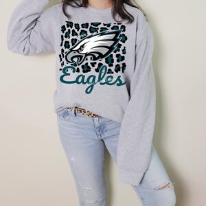 May include: A light grey sweatshirt with the Philadelphia Eagles logo. The design features a white and teal eagle head on a leopard print background, with the word "Eagles" in teal script. The sweatshirt is worn with light wash jeans.