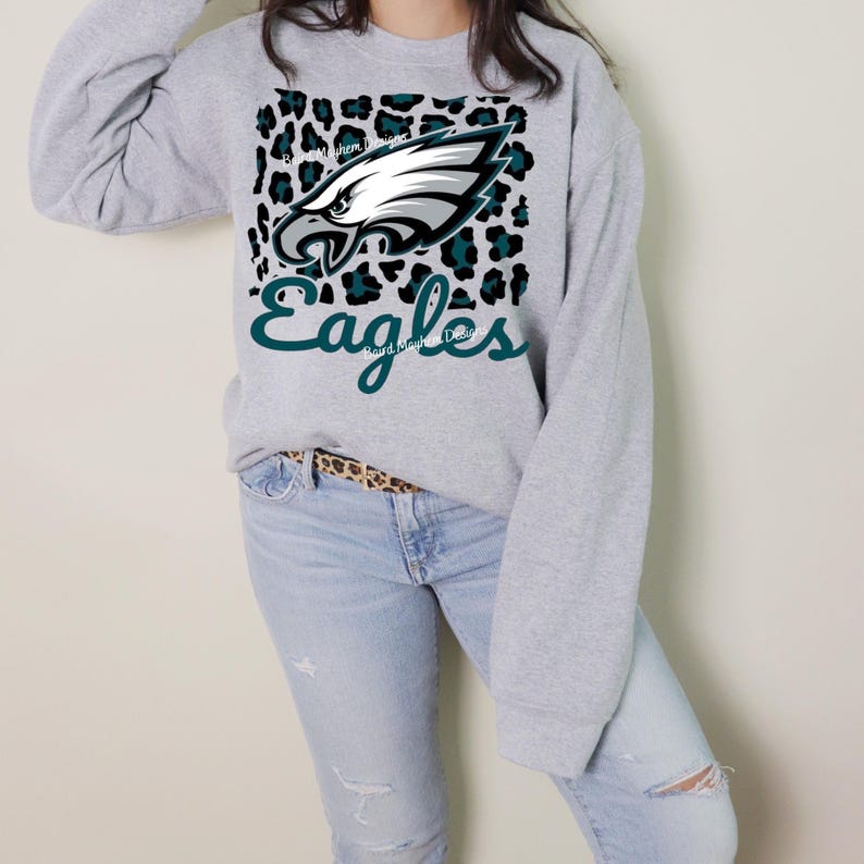 May include: A light grey sweatshirt with the Philadelphia Eagles logo. The design features a white and teal eagle head on a leopard print background, with the word "Eagles" in teal script. The sweatshirt is worn with light wash jeans.