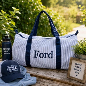 May include: A blue and white striped duffel bag with the name "Ford" embroidered in navy blue. A navy blue water bottle with "Adventure Awaits" text, a navy baseball cap with "Little Explorer" text, and a framed sign with "Work Hard Dream Big" text.