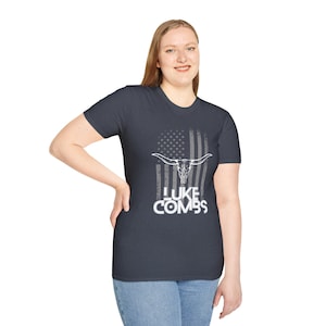 May include: Dark gray t-shirt featuring a white graphic of a longhorn skull over a faded American flag design. The text "LUKE COMBS" is printed in white below the flag. The shirt is worn by a person.