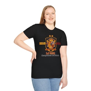 Può includere: Una t-shirt nera a maniche corte con una grafica dello stemma della nazionale di calcio spagnola sormontato da due stelle dorate. I numeri 2010 e 2026 fiancheggiano lo stemma, con la scritta La Roja Campeones Del Mundo in rosso e bianco.