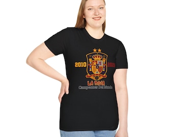 Może przedstawiać: Czarny t-shirt z krótkim rękawem z grafiką herbu hiszpańskiej reprezentacji narodowej w piłce nożnej z dwiema złotymi gwiazdami powyżej. Liczby 2010 i 2026 otaczają herb, a poniżej znajduje się napis La Roja Campeones Del Mundo w kolorze czerwonym i białym.