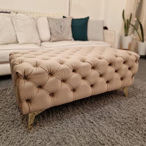 May include: A beige tufted ottoman with gold metal legs. The ottoman is sitting on a light brown shag rug.