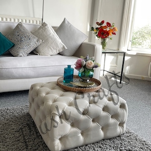 Puede incluir: Un otomano blanco capitoné con una bandeja tejida en la parte superior. El otomano está sentado en una alfombra gris en un ambiente de sala de estar. Hay almohadas en un sofá blanco en el fondo.