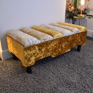 May include: A gold velvet storage ottoman with a tufted top and wooden legs. The ottoman is sitting on a gray carpet.