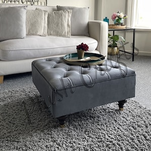 Puede incluir: Un otomano gris capitoné con cuatro patas de madera se encuentra sobre una alfombra de pelo gris frente a un sofá blanco con almohadas. El otomano está en un salón.