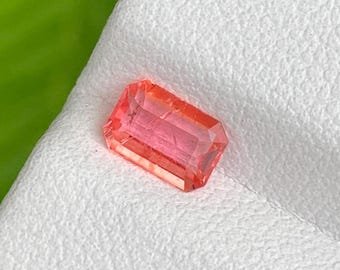Pode incluir: Uma pedra preciosa retangular e facetada com um tom vibrante de rosa-laranja. A gema está colocada sobre uma superfície branca texturizada, com um fundo verde desfocado. O corte da gema permite que a luz se reflita.
