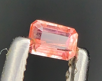 Peut inclure: Une pierre précieuse rectangulaire et facettée, de couleur pêche et rose, tenue par une pince argentée. La gemme a un aspect clair et transparent avec des inclusions internes. Le fond est gris foncé uni.