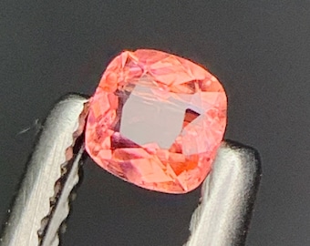 Peut inclure: Une pierre précieuse taillée en coussin, aux tons roses et orangés, est maintenue par une pince argentée. La gemme facettée a une surface réfléchissante, sur fond sombre.