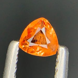 Puede incluir: Una gema naranja vibrante, de forma triangular, es sostenida por unas pinzas plateadas sobre un fondo oscuro. La gema tiene una superficie facetada, que capta la luz y muestra patrones internos. Esta gema es para joyería.