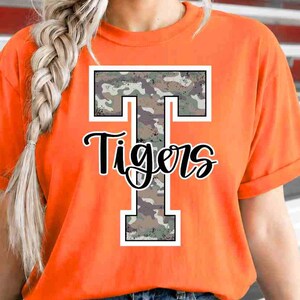 May include: An orange short-sleeved t-shirt featuring a large block letter T with a camouflage pattern and a white outline. The word Tigers is written in a black cursive script font across the centre of the letter T on the front of the shirt.