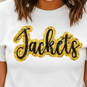 Pode incluir: Camiseta branca com a palavra "Jackets" em um design de glitter dourado. A palavra é delineada em preto. A camiseta é de gola redonda.