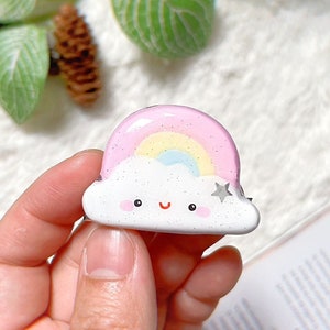 Op de afbeelding: Een klein keramiek beeldje van een witte wolk met een roze en gele regenboog erop. De wolk heeft een vrolijk gezicht en een kleine zilveren ster aan de zijkant.