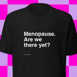 May include: Black t-shirt with the text "Menopause. Are we there yet?" in white. The shirt has a crew neck and short sleeves. The text is centered on the front of the shirt. The background is a checkered pattern of pink and purple squares.